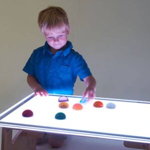 Jongen in blauwe shirt speelt met kleurrijke vormen op een verlichte lichttafel XL.
