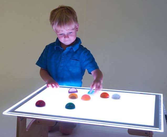 Jongen in blauwe shirt speelt met kleurrijke vormen op een verlichte lichttafel XL.