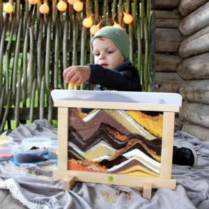 Kind met zand schilderij, strooit kleurrijk zand op een plakvlak, omgeven door potjes met zand en een houten achtergrond.