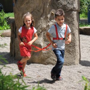 Rood paardentuigje voor kinderen, gedragen door een meisje en een jongen, spelend in een buitenspeelomgeving.
