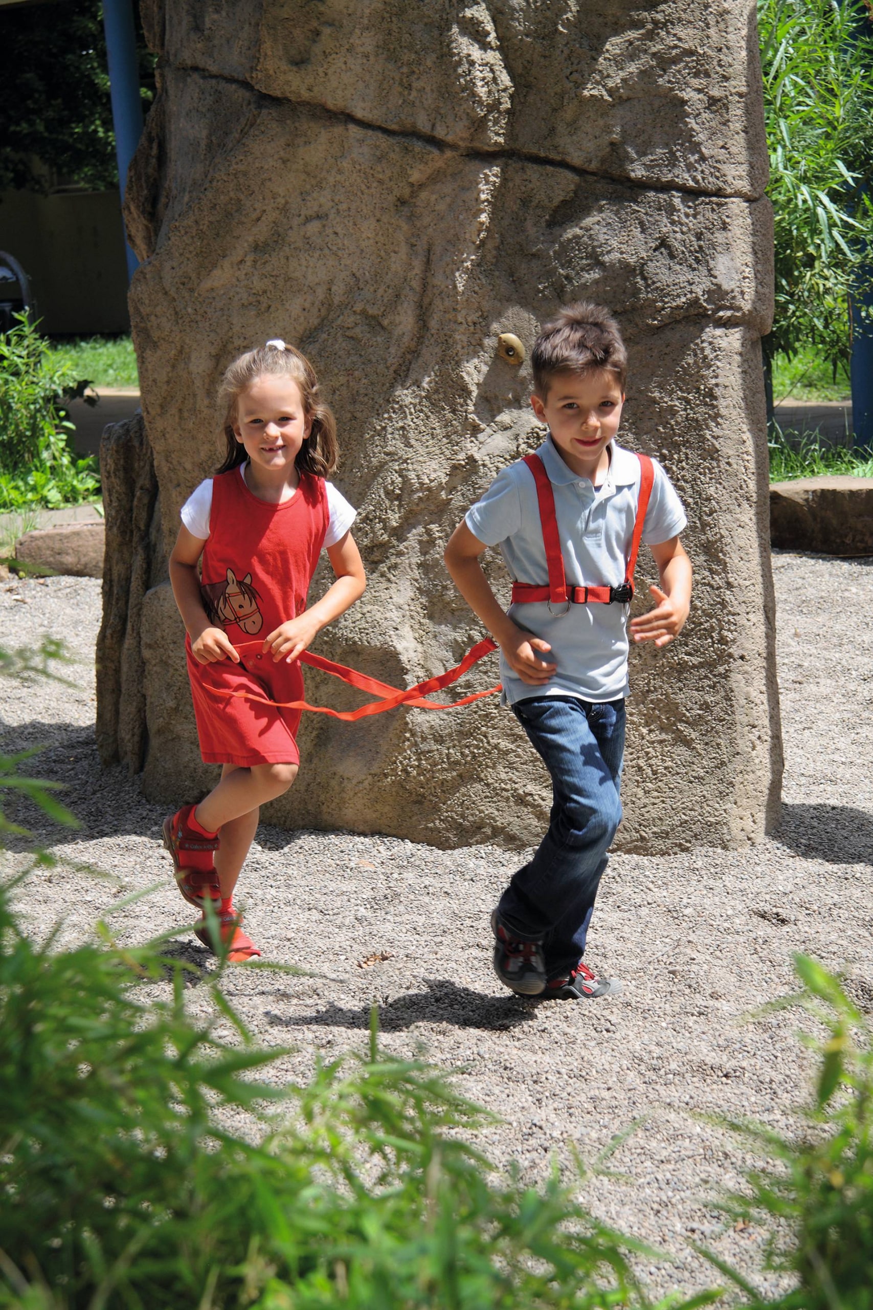 Rood paardentuigje voor kinderen, gedragen door een meisje en een jongen, spelend in een buitenspeelomgeving.
