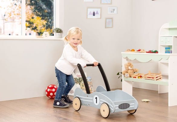 Zilveren loopwagen in de vorm van een sportauto, met een meisje dat er vrolijk mee speelt in een speelse omgeving.