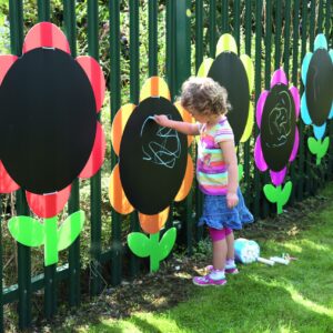 Vrolijke bloemborden in bloemdessin, met een kind dat op een zwart bord tekent, tegen een groene schutting.
