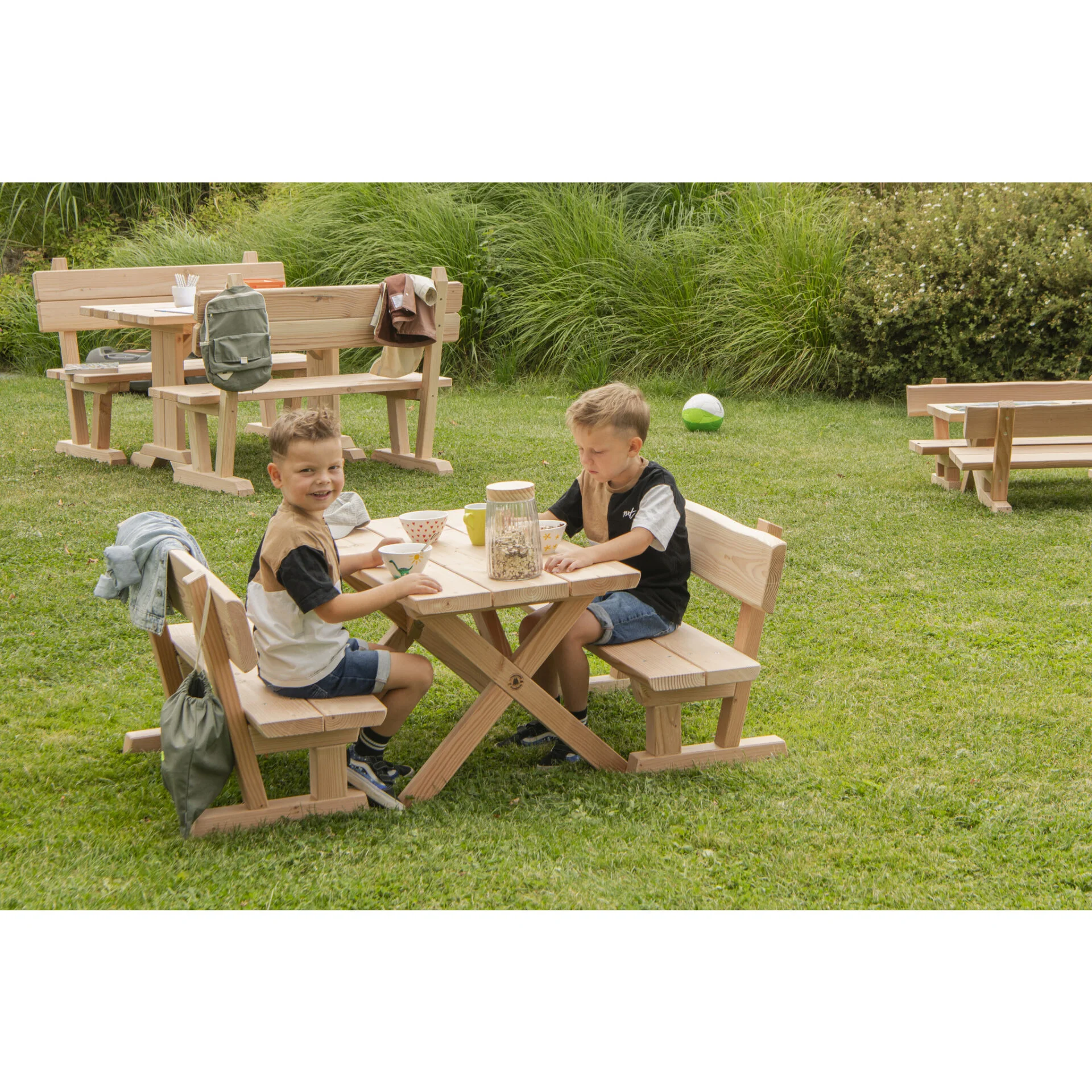 Kinderen zitten aan een houten buitentafel met banken, spelen en snacken in een groene tuin.