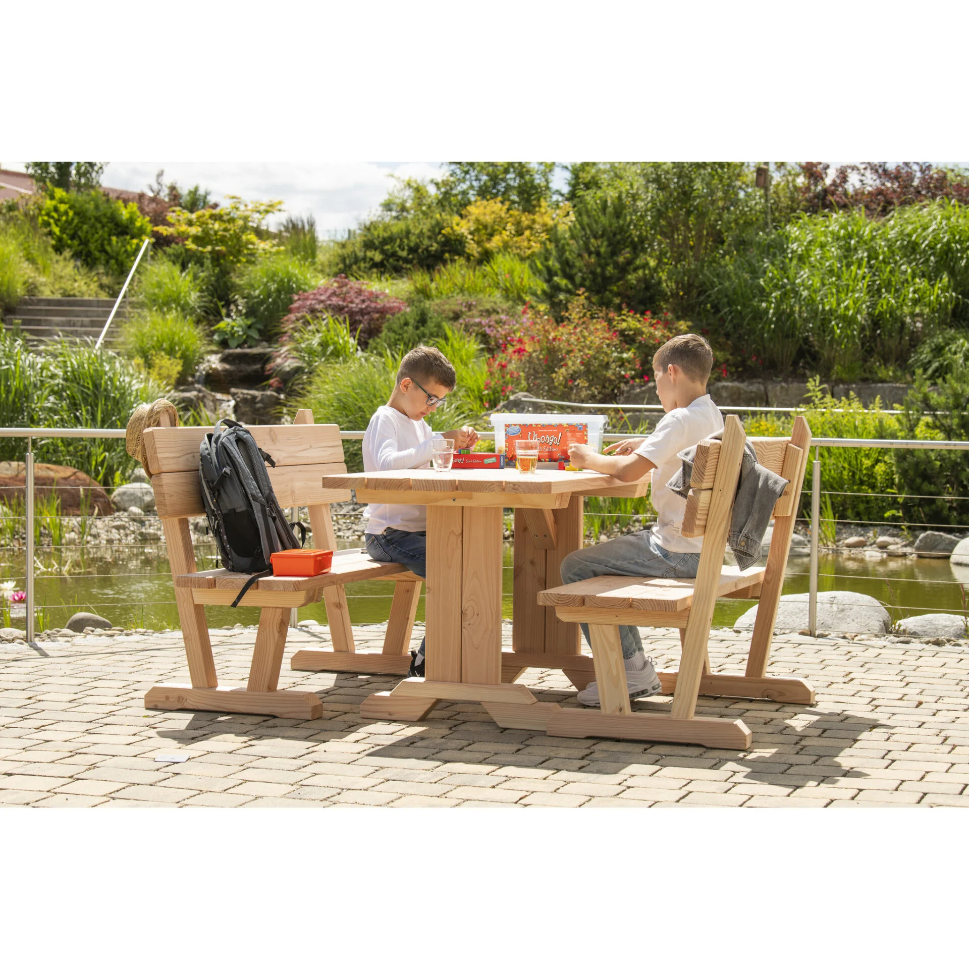 Twee kinderen zitten aan een houten buitentafel met stoelen, omringd door groen en waterpartijen.