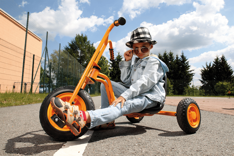 Stoere Chopper driewieler in fel oranje, met lage zitting en hoog stuur, gebruikt door een kind in een spijkeroutfit.