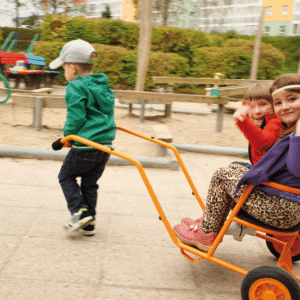 Kinderen spelen met een oranje riksja in een speeltuin, één zit erin en de ander duwt.