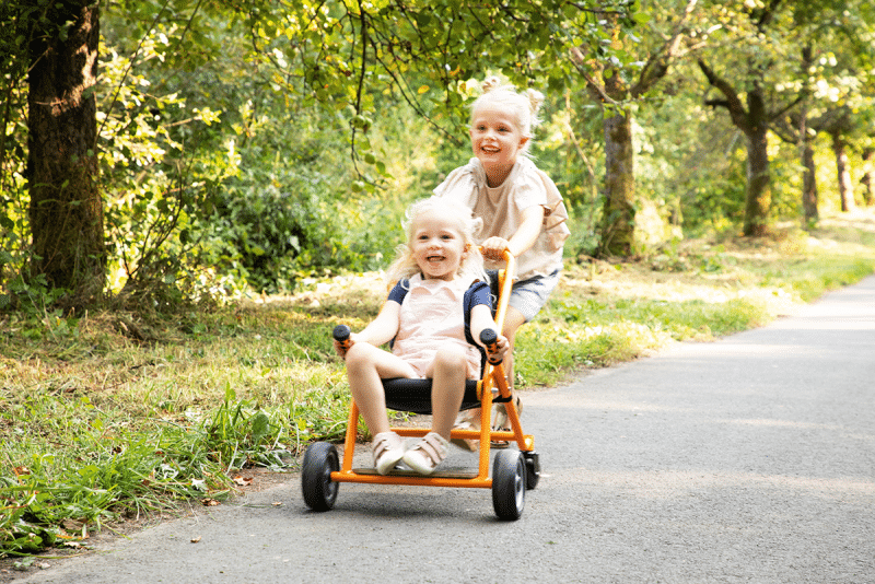 Kinderen duwen elkaar in een oranje Toffe Buggy met zwenkwielen, ideaal voor buiten spelen en het vervoeren van knuffels.