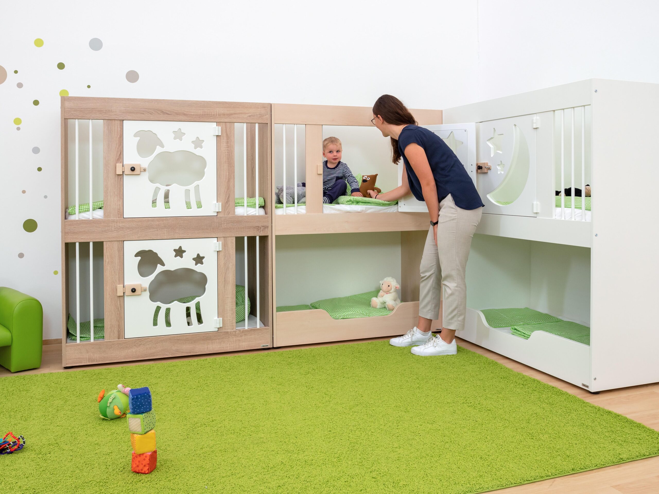 Stapelbed voor kinderen met één deur, houten afwerking, en een kind dat speelt met een volwassene. Groene matten en spee...