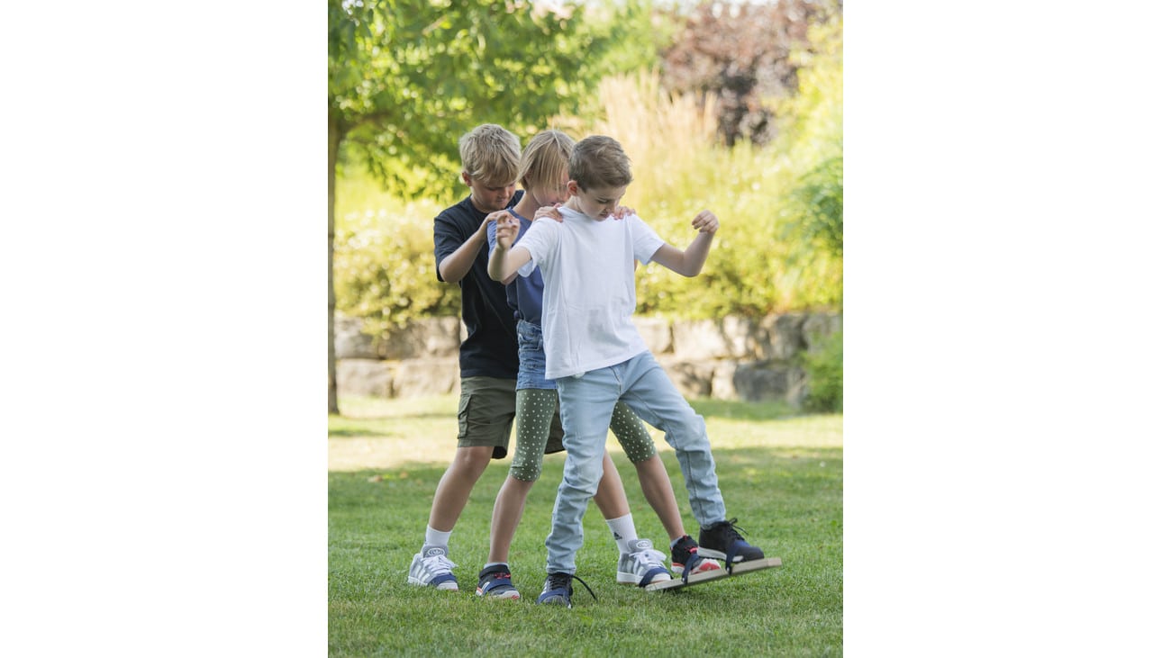 Drie kinderen spelen samen op zomerskis op gras, lachend en in beweging, met een groene achtergrond.