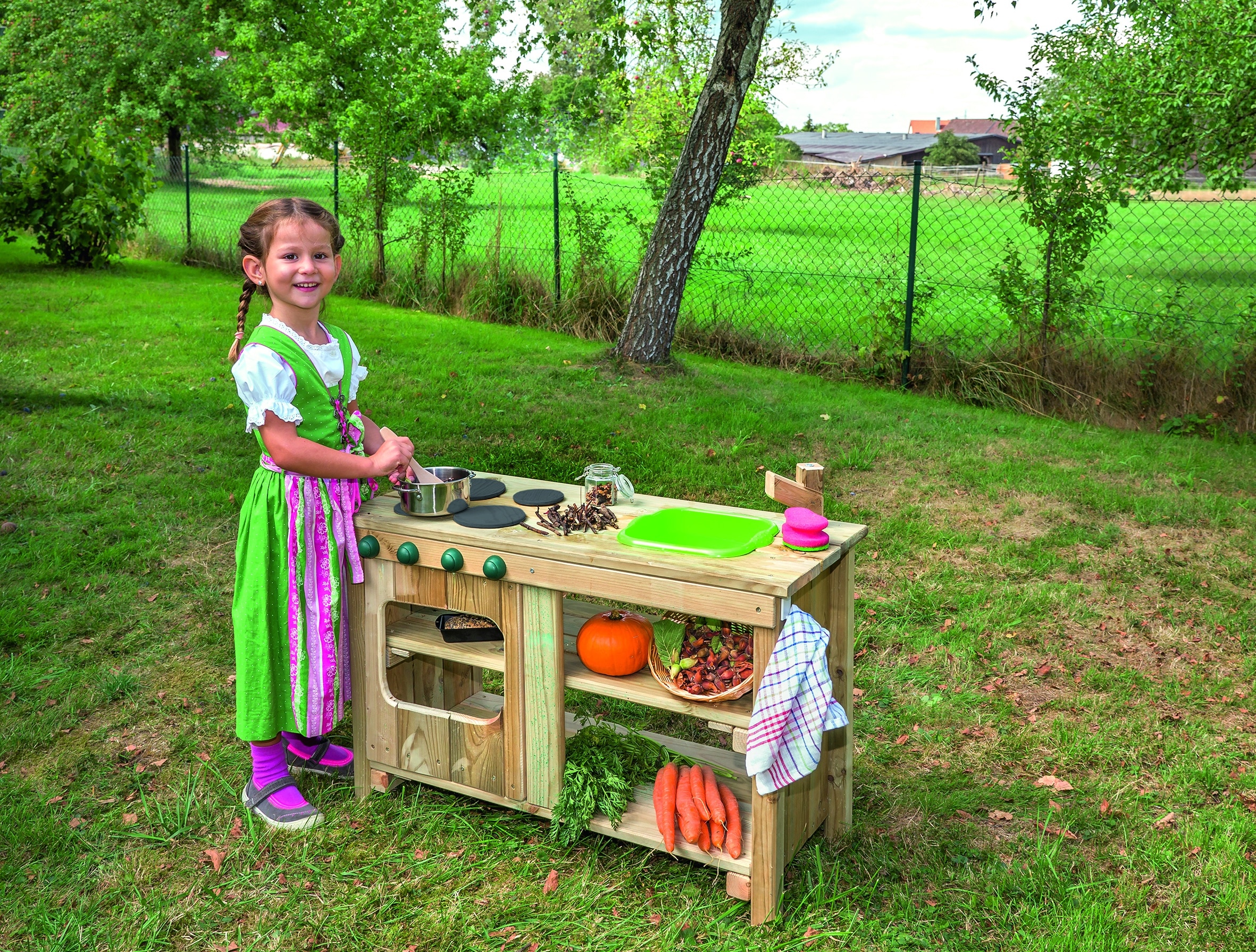 Buiten keuken voor kleuters met speeltoestellen, groenten en een kind dat aan het koken is in een groene omgeving.