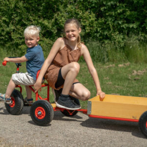 Kleine ROSE driewieler met ballonwielen, houten zitting en aanhanger, gebruikt door twee kinderen in een groene omgeving.