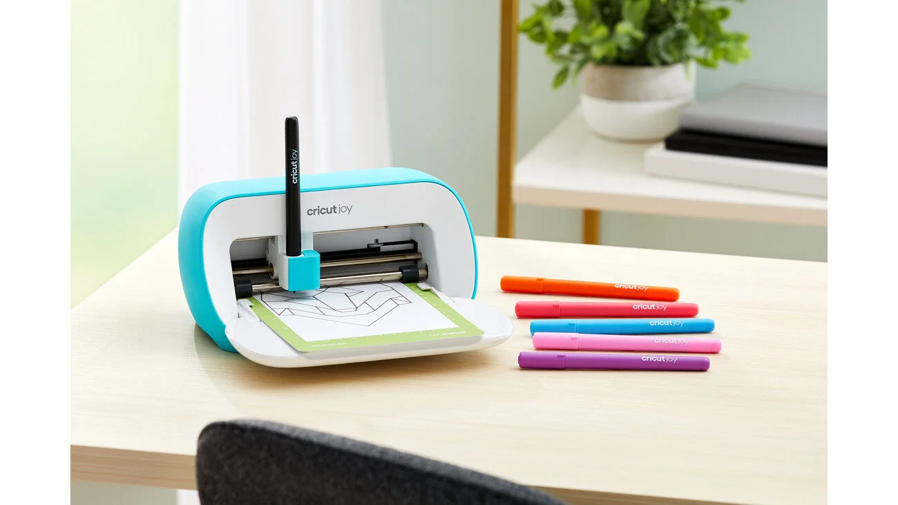Cricut Joy snijplotter in turquoise met een groene snijmat en kleurrijke pennen op een houten tafel.