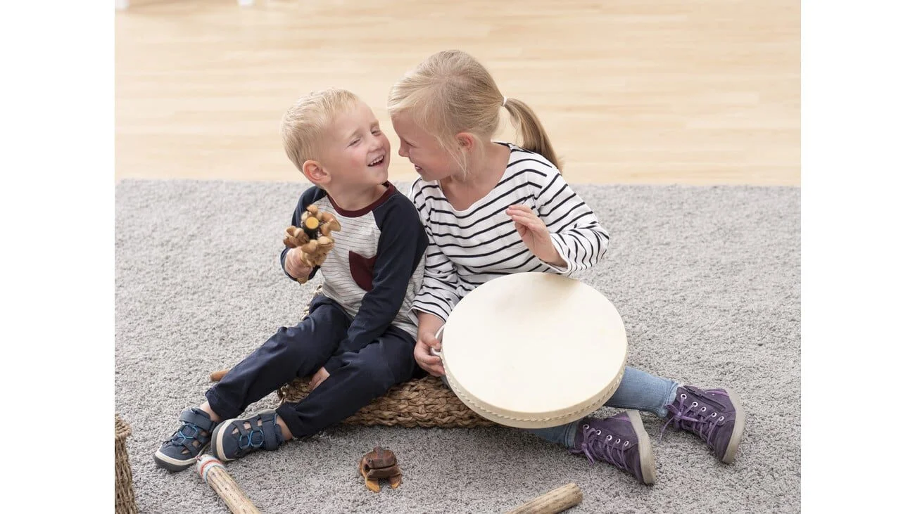 Kinderen zitten op de grond met muziekinstrumenten, waaronder een trommel en kikker-guiros.