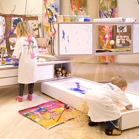 Creatieve speelruimte met kinderen die schilderen, kleurrijke kunstwerken en speelmateriaal zichtbaar.
