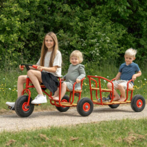 Taxi-aanhanger met ballonwielen, rood, met twee zitplaatsen, bevestigd aan een driewieler, kinderen rijden op een pad.