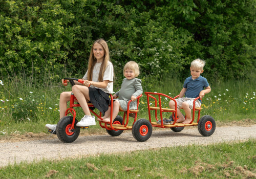 Taxi-aanhanger met ballonwielen, rood, met twee zitplaatsen, bevestigd aan een driewieler, kinderen rijden op een pad.