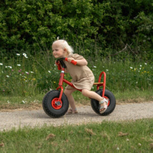 ROZE loopfiets met ballonbanden, houten zitting en metalen frame, gebruikt door een kind op een pad in de natuur.