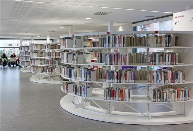 Moderne bibliotheekkasten in ronde vorm, gevuld met boeken, in een lichte, open ruimte met studeertafels.